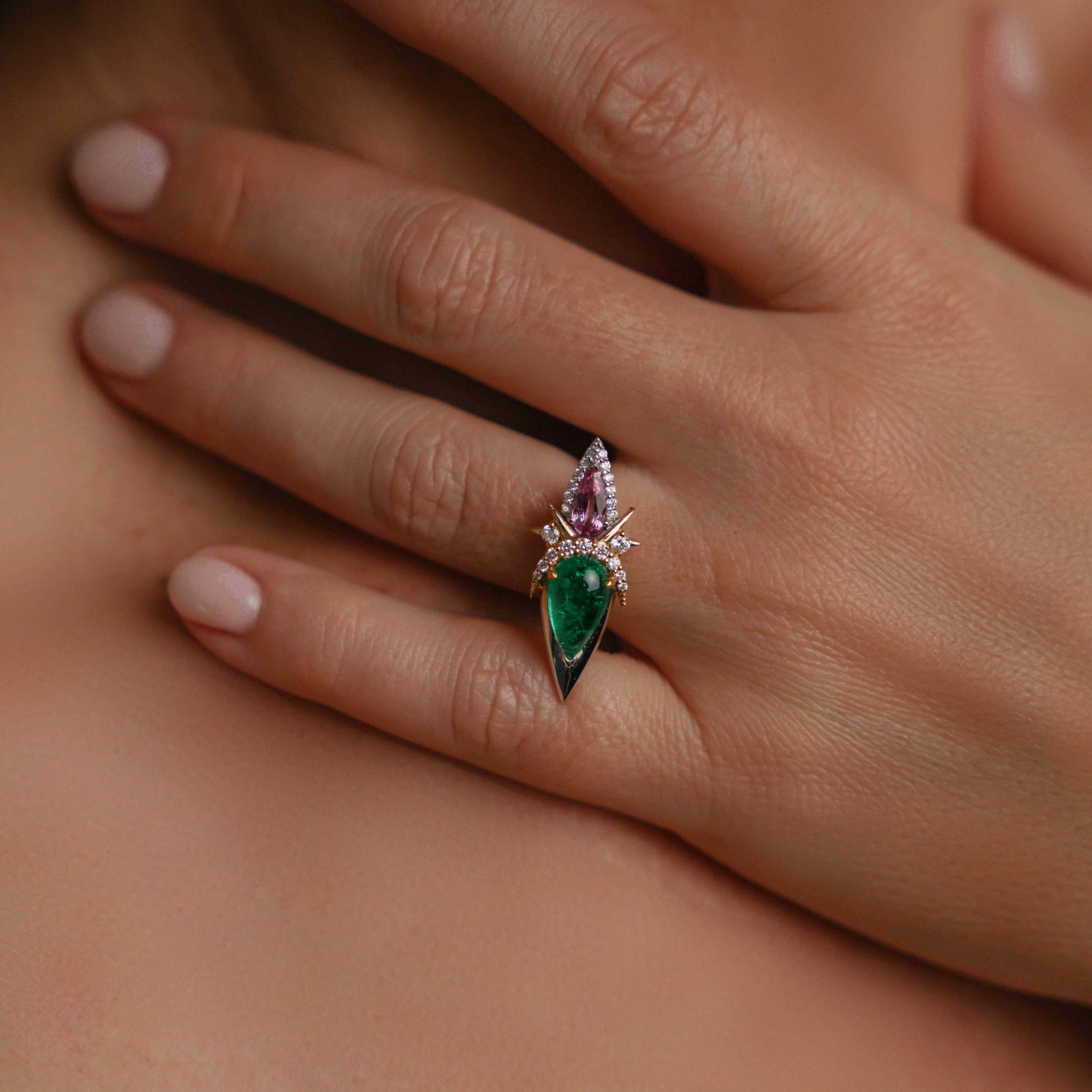 Model wearing elegant ring with a large green emerald, a pink sapphire, and diamond accents on a gold band on engagement ring finger.