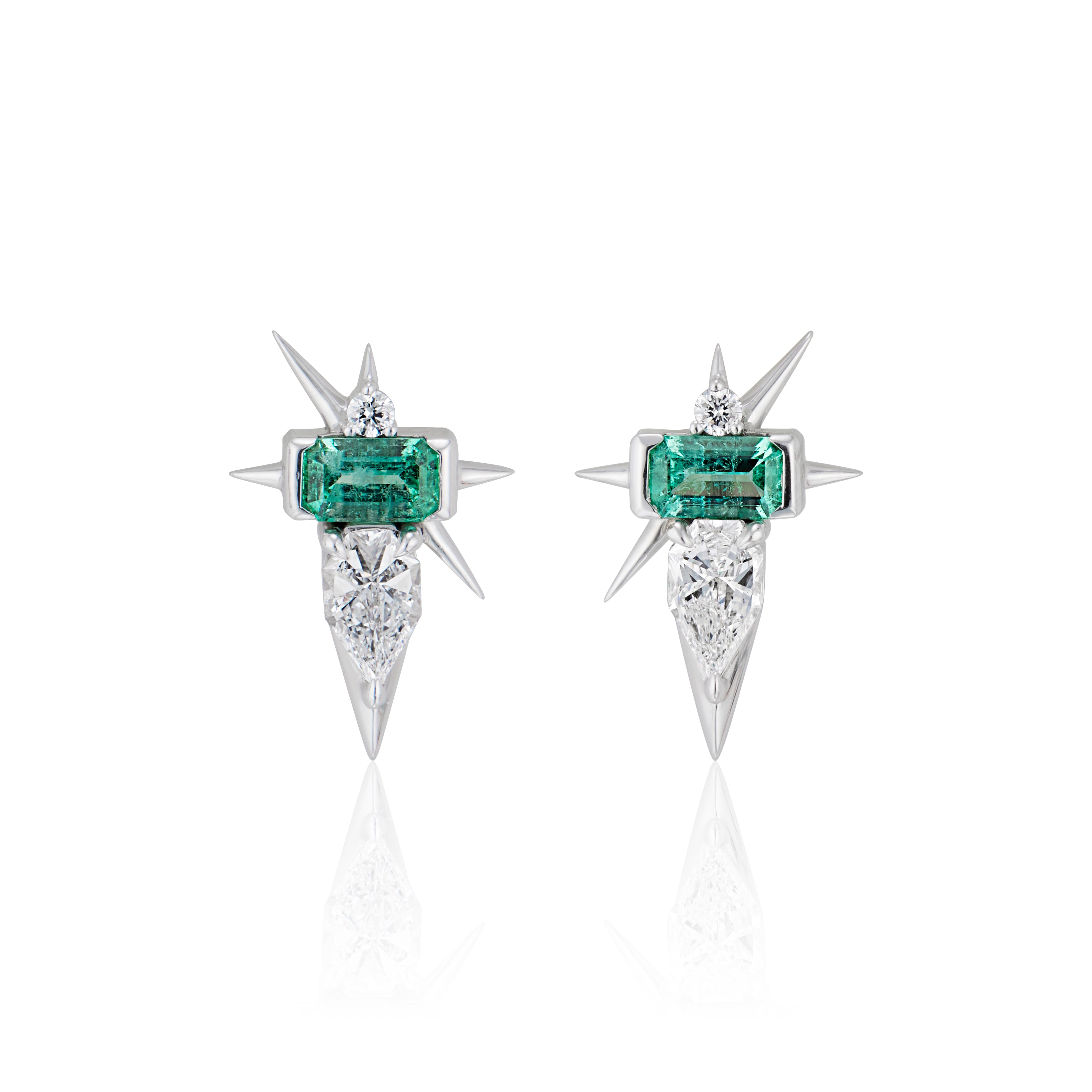 Emerald and diamond starburst earrings on a white reflective surface.