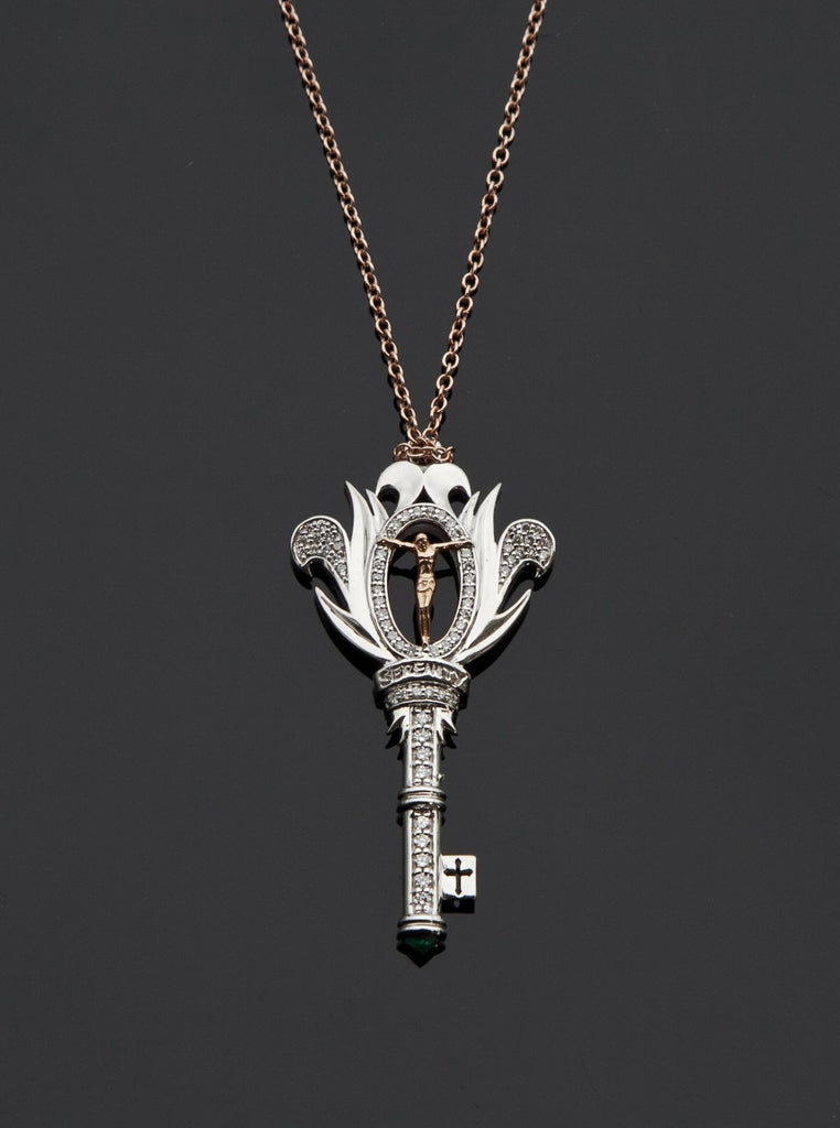 Bespoke key-shaped necklace in white gold with rose gold crucifix stretching across an oval diamond outlined cutout. The head of the key resembles a side view of an opening flower with white gold and diamond accented petals. Below the crucifix, the engraved word “Serenity”. The key’s stem has a single line of round diamonds with a white gold collar in the middle. An emerald is at the tip of the key and a single bit with a cross cutout extends out. The charm hangs from a rose gold cable chain.