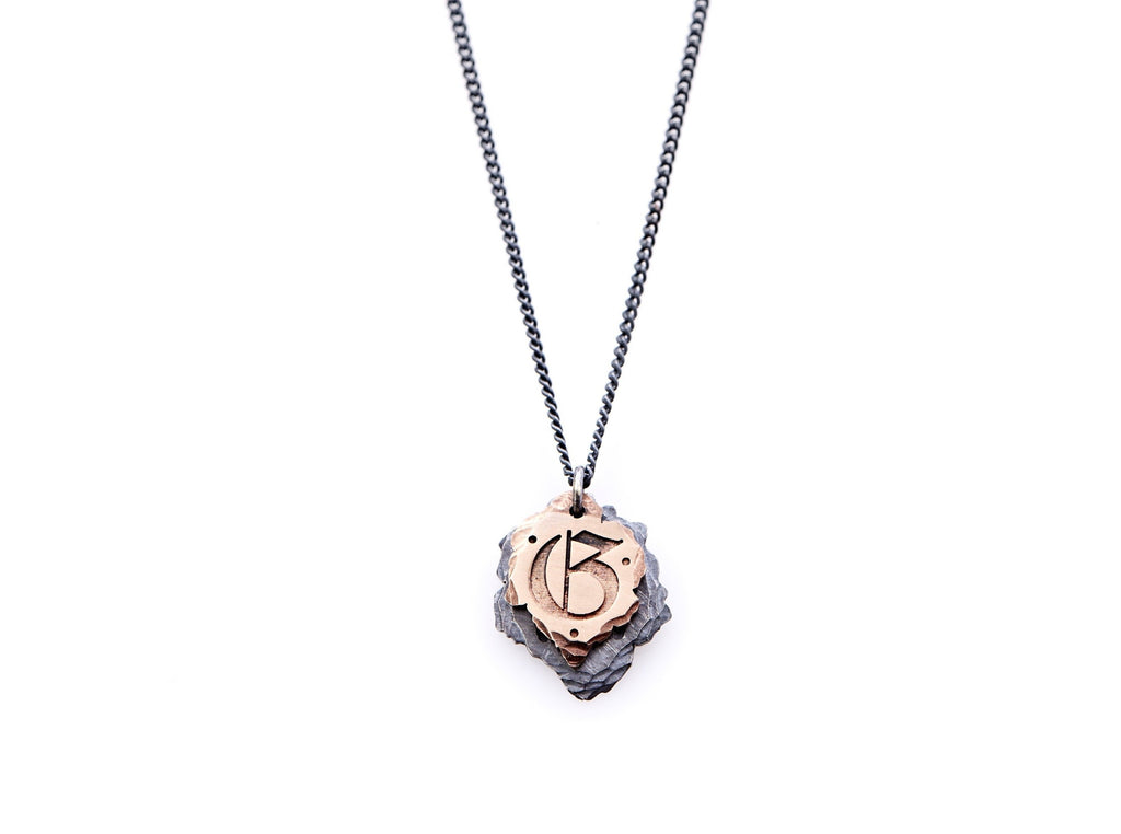 Hand crafted, customizable Initial Necklace with 2 jagged-edged plates hanging from an oxidized sterling silver curbed chain. Top plate is hand forged rose gold with a custom engraving of the letter "G" in Old English lettering with 2 hand engraved dots on either side. Bottom plate is hand forged oxidized sterling silver and slightly larger than the rose gold plate. The top plate partially covers the bottom plate, and edges of cross- bones are visible on the bottom plate.