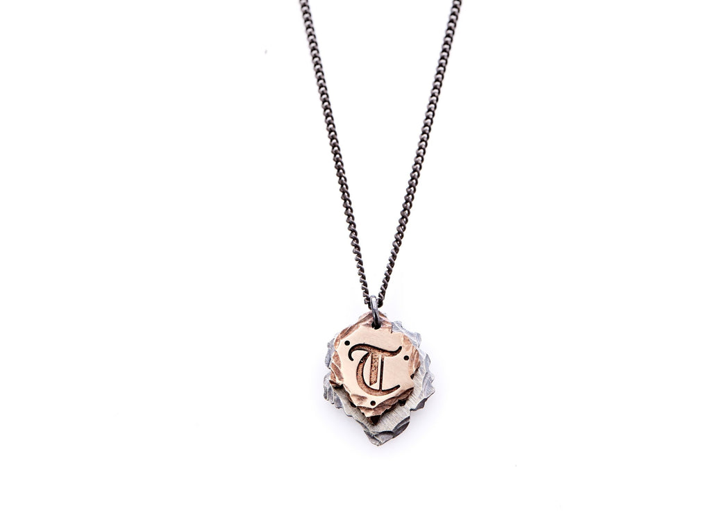Hand crafted, customizable Initial Necklace with 2 jagged-edged plates hanging from an oxidized sterling silver curbed chain. Top plate is hand forged rose gold with a custom engraving of the letter "T" in Old English lettering with 2 hand engraved dots on either side. Bottom plate is hand forged oxidized sterling silver and slightly larger than the rose gold plate. The top plate partially covers the bottom plate, and edges of cross- bones are visible on the bottom plate.