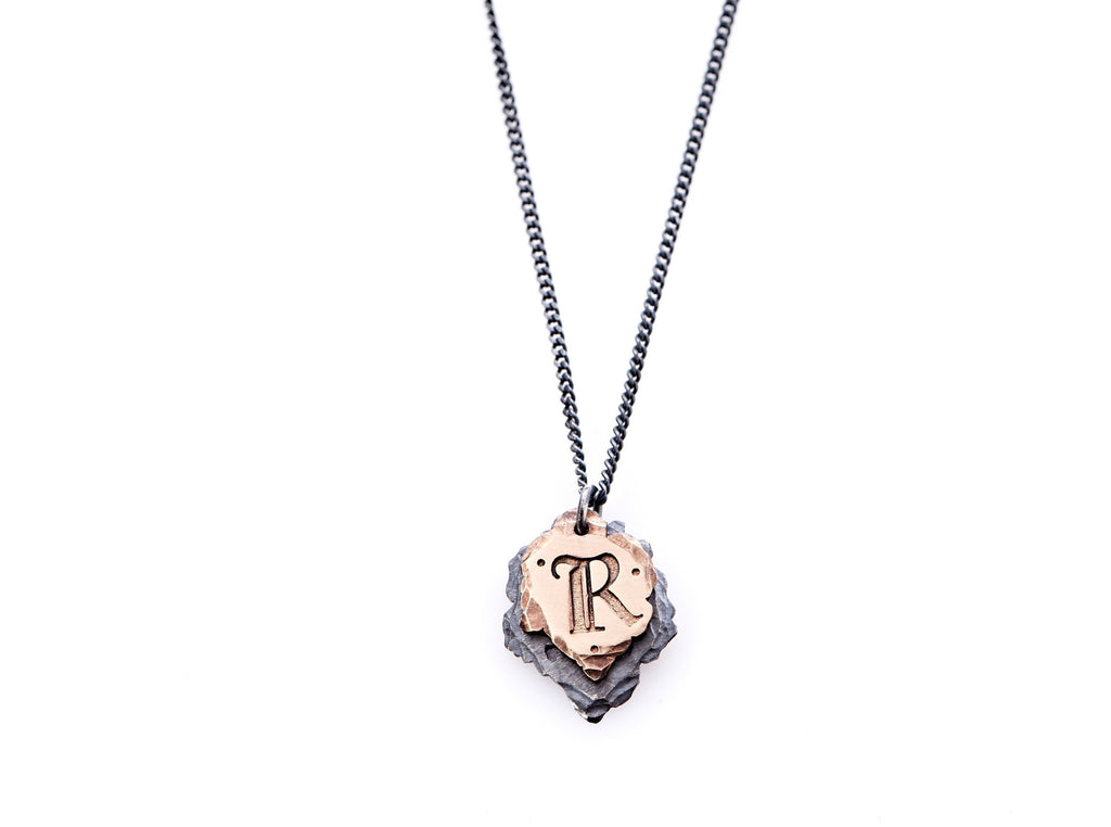 Hand crafted, customizable Initial Necklace with 2 jagged-edged plates hanging from an oxidized sterling silver curbed chain. Top plate is hand forged rose gold with a custom engraving of the letter "R" in Old English lettering with 2 hand engraved dots on either side. Bottom plate is hand forged oxidized sterling silver and slightly larger than the rose gold plate. The top plate partially covers the bottom plate, and edges of cross- bones are visible on the bottom plate.