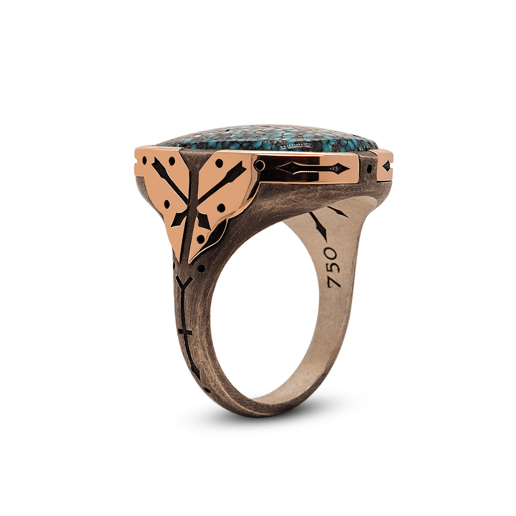  Side angle of the men’s turquoise ring standing up on its base. Handset turquoise stone juts slightly out of the setting and forms a rounded dome in the center. Rose gold setting wraps over the shoulder of the ring. The polished rose gold contrasts with the oxidized sterling silver band, complimenting the turquoise stone. The bottom of the setting is visible and has an engraved dot on the sterling silver tab between the rose gold accents.