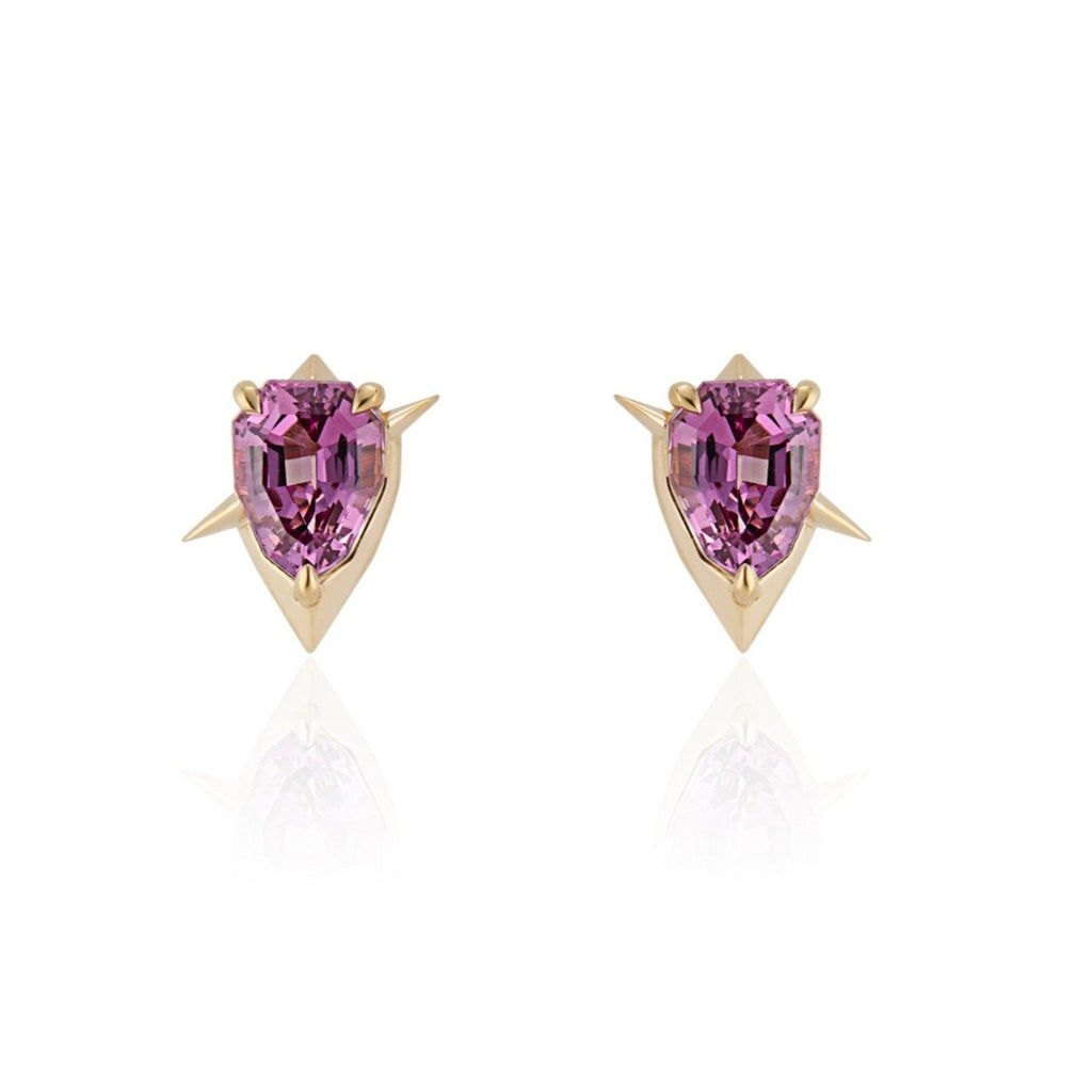 Front view. Shield-cut pink garnet stud earrings in a diamond-shaped yellow gold setting. 2 short yellow gold spikes extend out diagonally in either direction, mirroring each other, as if to pass through the setting like a sword.