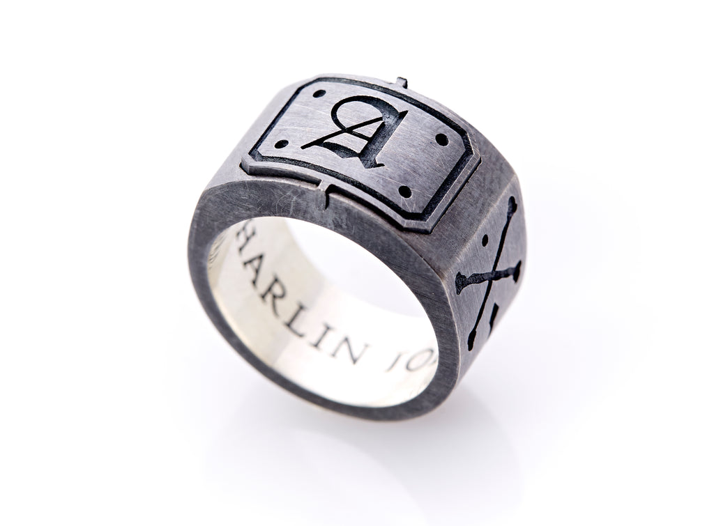 Text: Side view of a bespoke jewelry design, the signet ring, balanced on its base. The A insignia is at the center of the head of the ring which is slightly thicker and less convex. Black engraved text on the interior polished sterling silver base of the band is visible which reads, “Harlin Jones.”