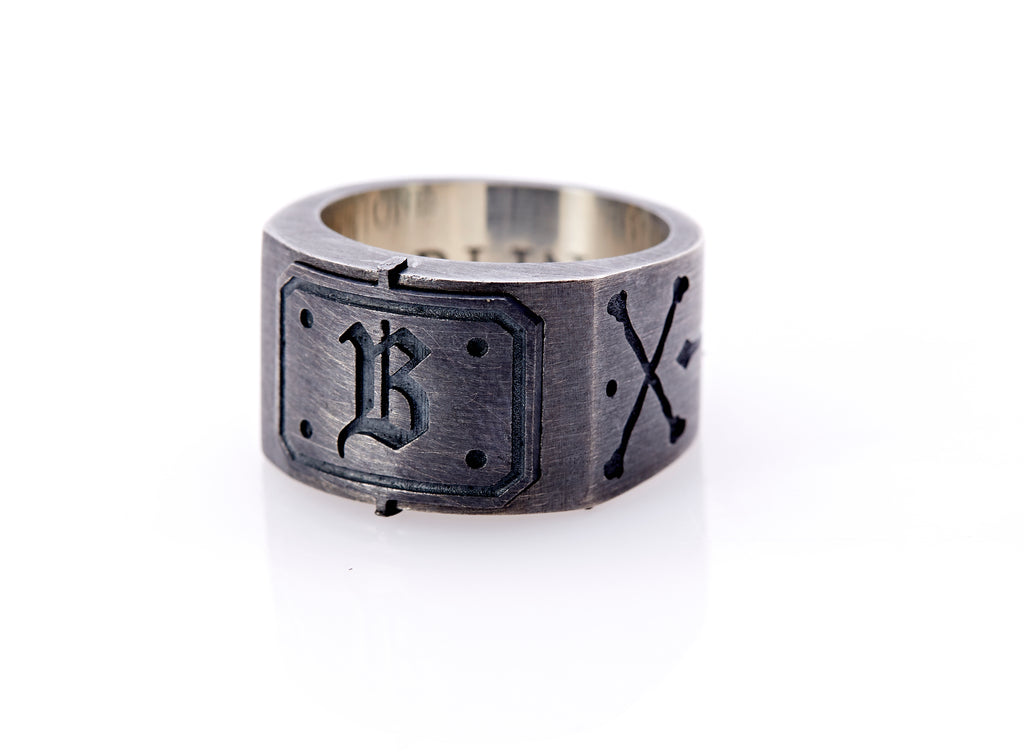 Oxidized sterling silver men’s signet ring with a thick flat band hand-crafted in New York. Semi-raised octagonal engraved top cap with a customizable insignia design of an engraved letter “B” in Old English Style lettering and 4 engraved dots at each corner. The initial on the ring is surrounded by a bold black engraved border that follows the inside edge of the rectangle. The rectangle’s top and bottom edge is accented by a small lip in the center that wraps around the corner edge of the ring.