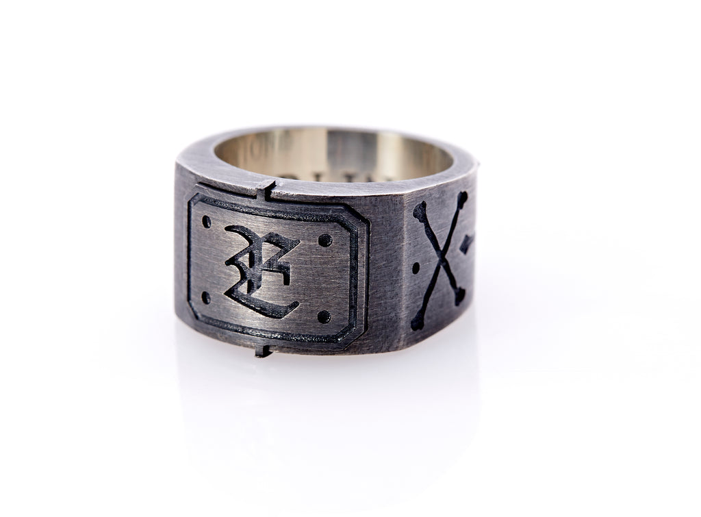 Oxidized sterling silver men’s signet ring with a thick flat band hand-crafted in New York. Semi-raised octagonal engraved top cap with a customizable insignia design of an engraved letter “E” in Old English Style lettering and 4 engraved dots at each corner. The initial on the ring is surrounded by a bold black engraved border that follows the inside edge of the rectangle. The rectangle’s top and bottom edge is accented by a small lip in the center that wraps around the corner edge of the ring.