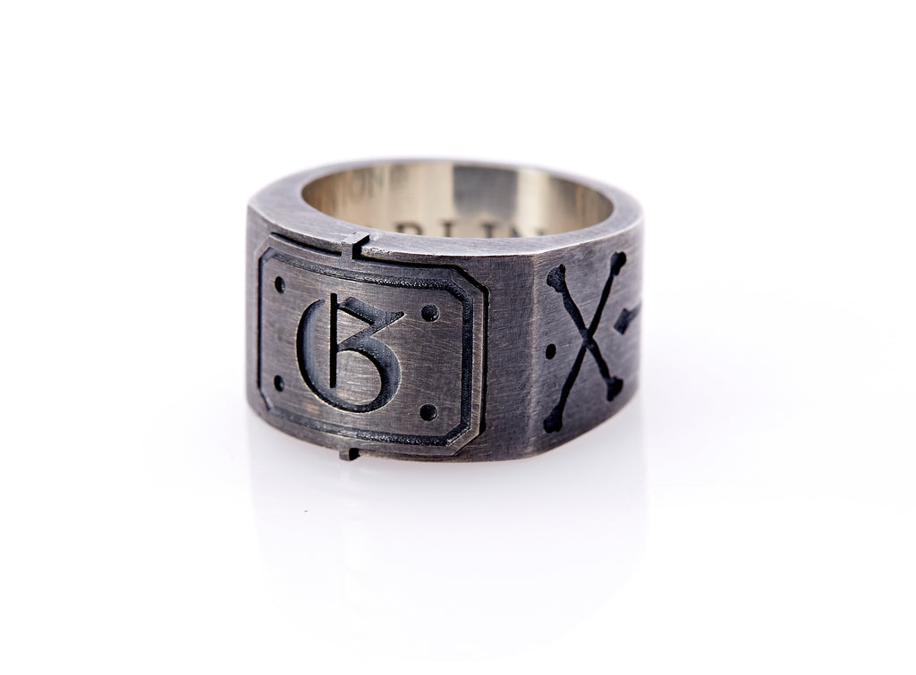Oxidized sterling silver men’s signet ring with a thick flat band hand-crafted in New York. Semi-raised octagonal engraved top cap with a customizable insignia design of an engraved letter “G” in Old English Style lettering and 4 engraved dots at each corner. The initial on the ring is surrounded by a bold black engraved border that follows the inside edge of the rectangle. The rectangle’s top and bottom edge is accented by a small lip in the center that wraps around the corner edge of the ring.