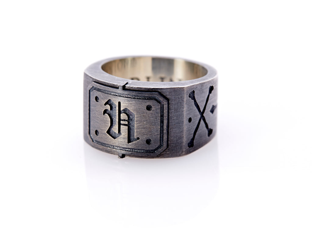 Oxidized sterling silver men’s signet ring with a thick flat band hand-crafted in New York. Semi-raised octagonal engraved top cap with a customizable insignia design of an engraved letter “H” in Old English Style lettering and 4 engraved dots at each corner. The initial on the ring is surrounded by a bold black engraved border that follows the inside edge of the rectangle. The rectangle’s top and bottom edge is accented by a small lip in the center that wraps around the corner edge of the ring.