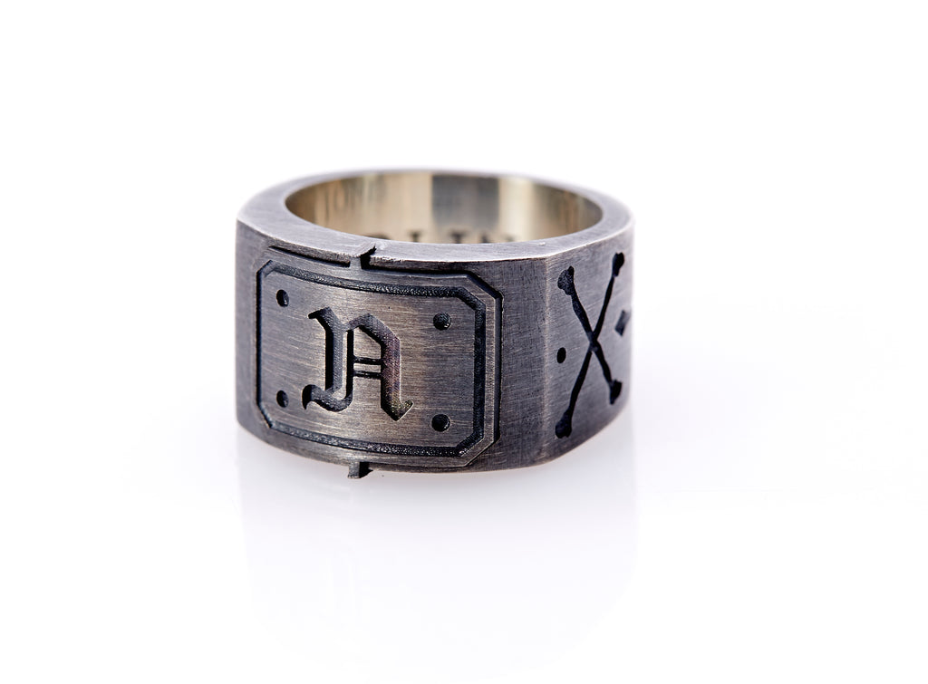 Oxidized sterling silver men’s signet ring with a thick flat band hand-crafted in New York. Semi-raised octagonal engraved top cap with a customizable insignia design of an engraved letter “N” in Old English Style lettering and 4 engraved dots at each corner. The initial on the ring is surrounded by a bold black engraved border that follows the inside edge of the rectangle. The rectangle’s top and bottom edge is accented by a small lip in the center that wraps around the corner edge of the ring.
