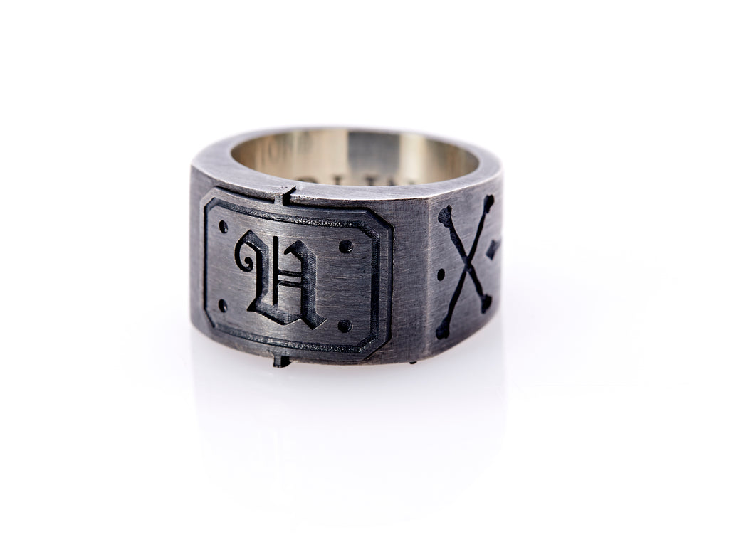 Oxidized sterling silver men’s signet ring with a thick flat band hand-crafted in New York. Semi-raised octagonal engraved top cap with a customizable insignia design of an engraved letter “U” in Old English Style lettering and 4 engraved dots at each corner. The initial on the ring is surrounded by a bold black engraved border that follows the inside edge of the rectangle. The rectangle’s top and bottom edge is accented by a small lip in the center that wraps around the corner edge of the ring.