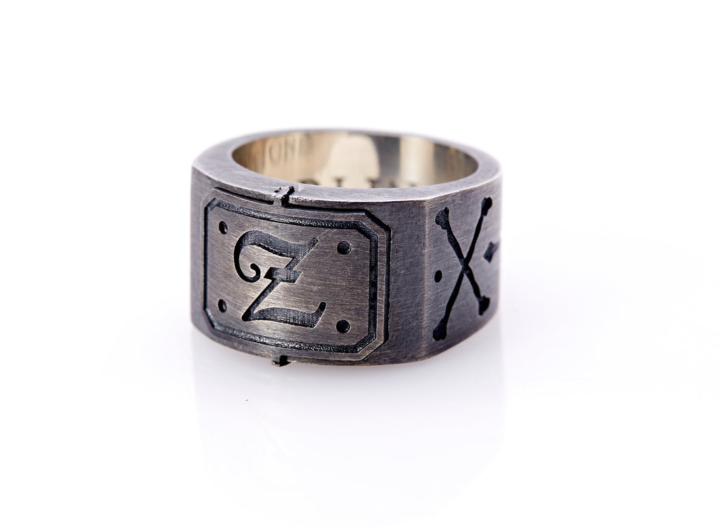 Oxidized sterling silver men’s signet ring with a thick flat band hand-crafted in New York. Semi-raised octagonal engraved top cap with a customizable insignia design of an engraved letter “Z” in Old English Style lettering and 4 engraved dots at each corner. The initial on the ring is surrounded by a bold black engraved border that follows the inside edge of the rectangle. The rectangle’s top and bottom edge is accented by a small lip in the center that wraps around the corner edge of the ring.
