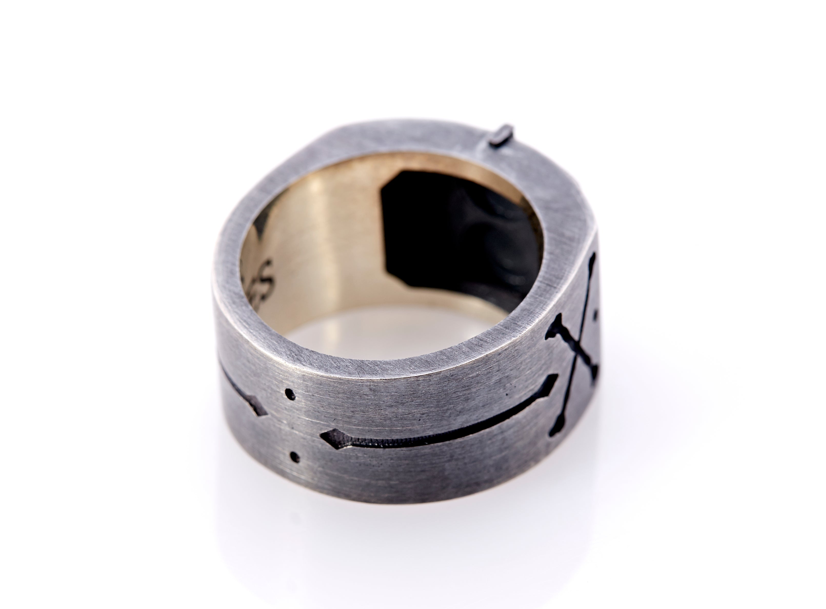 Side and base of signet ring band with unique black hand-engraved details, including arrows, dots and cross-bones. The center of the base of the outer band has 2 vertical dots. Continuing towards the head of the ring in the lower shank section, is an elongated, centered two-way arrow. This leads to cross-bones and a centered dot in the upper shank. The opposite side mirrors this hand-crafted design. The underside of the semi-raised octagonal rectangle is black.