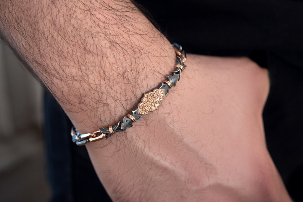 Closeup on man’s wrist wearing the Harlin Jones oxidized sterling silver and rose gold bracelet. The filigree plate sits in the center of the wrist and is roughly the size of a Tic Tac. As the arm points downwards, the bracelet falls to where the wrist meets the hand. The bracelet is the width of a thick rubber band.