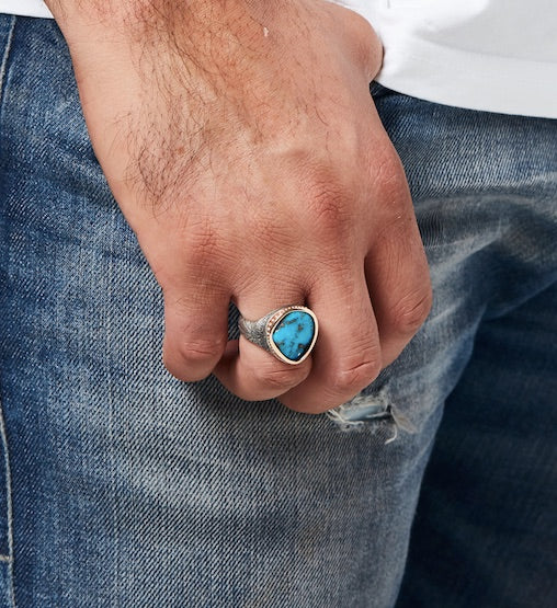 Closeup on man’s hand wearing the bespoke Morenci Turquoise Ring on his ring finger. Thin band at lower shank flares out to double its size at the upper shank, fitting snugly around the base of the knuckle. The ring’s stone and rose gold setting extends from the base of the knuckle to just below the first joint. The stone has a tear-drop shape that sits on the top of the finger, leaving the side of the ring’s band and its Thunderbird engraving visible.