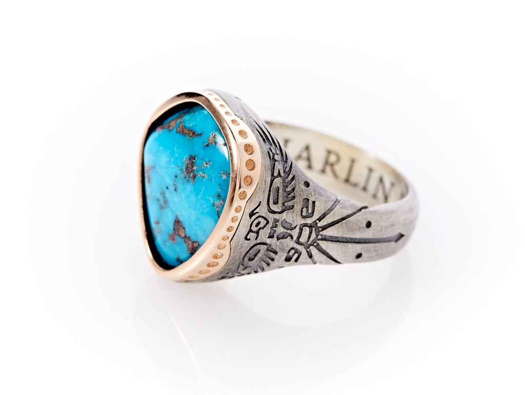Side view of Turquoise Thunderbird Ring with rose gold petal-shaped bezel and oxidized silver band. Just below the setting, a hand-crafted Thunderbird design engraved into the sterling silver metal. Its wings reach the width of the ring’s setting and its tail extends the length of the band. A cutting of Morenci turquoise with iron pyrite is handset, creating a unique, natural shape. On the interior of the band, the engraved word “Harlin”.