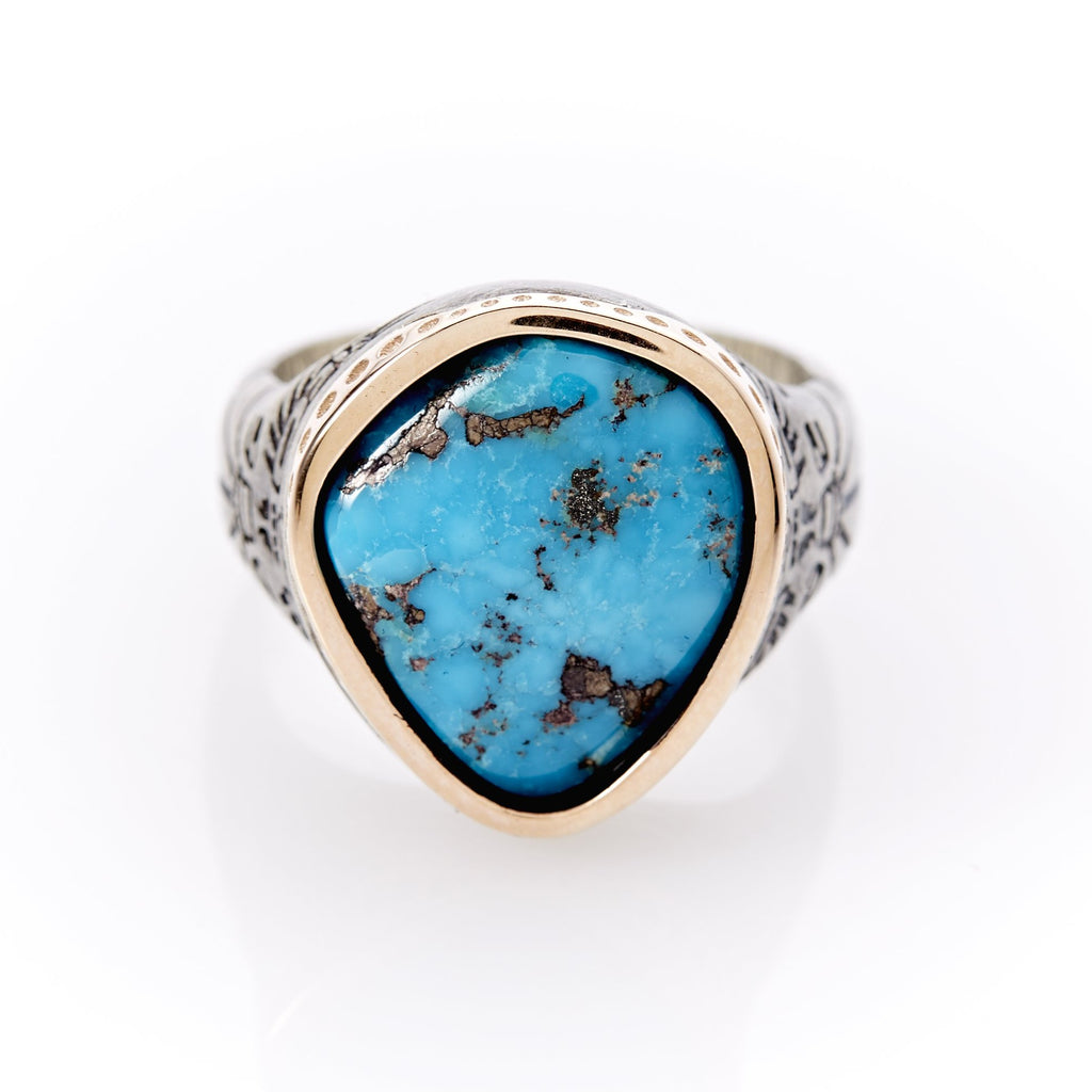 Men’s turquoise ring with a rose gold bezel setting constructed in a teardrop shape. Top of teardrop on band is accentuated by small, circular-shaped depressions. Inside the setting is a unique, organic cutting of turquoise with predominantly blue coloring and flecks of iron pyrite. The ring’s band consists of sterling silver with hand- crafted engraved designs cut into it.