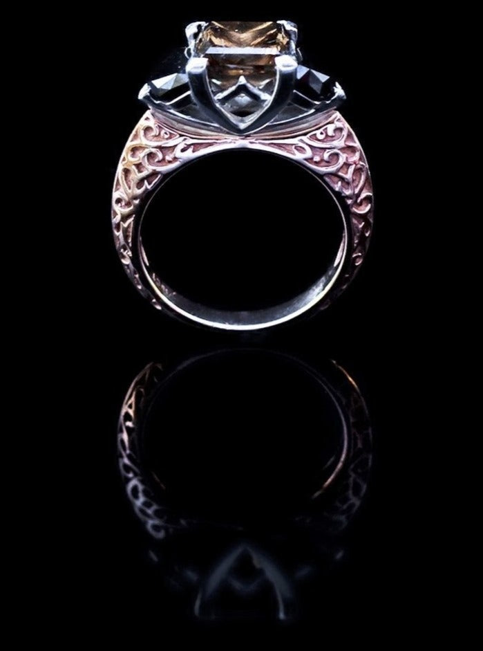  Side angle of bespoke ring by a New York jewelry designer with Cognac diamond center stone flanked by 2 black diamonds. The stones are held in place by a white gold setting and prongs. The setting rests on a white gold band with a white and rose gold filigree design embellishment. The band is thick at the shoulders and gradually tapers down into a thinner base.