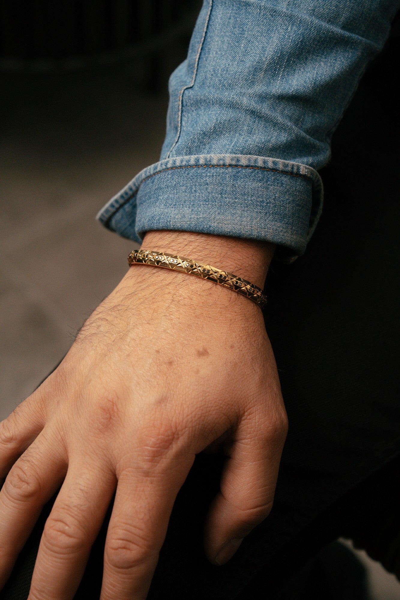 Hand wearing a gold bracelet with rolled-up jeans sleeve on a dark background