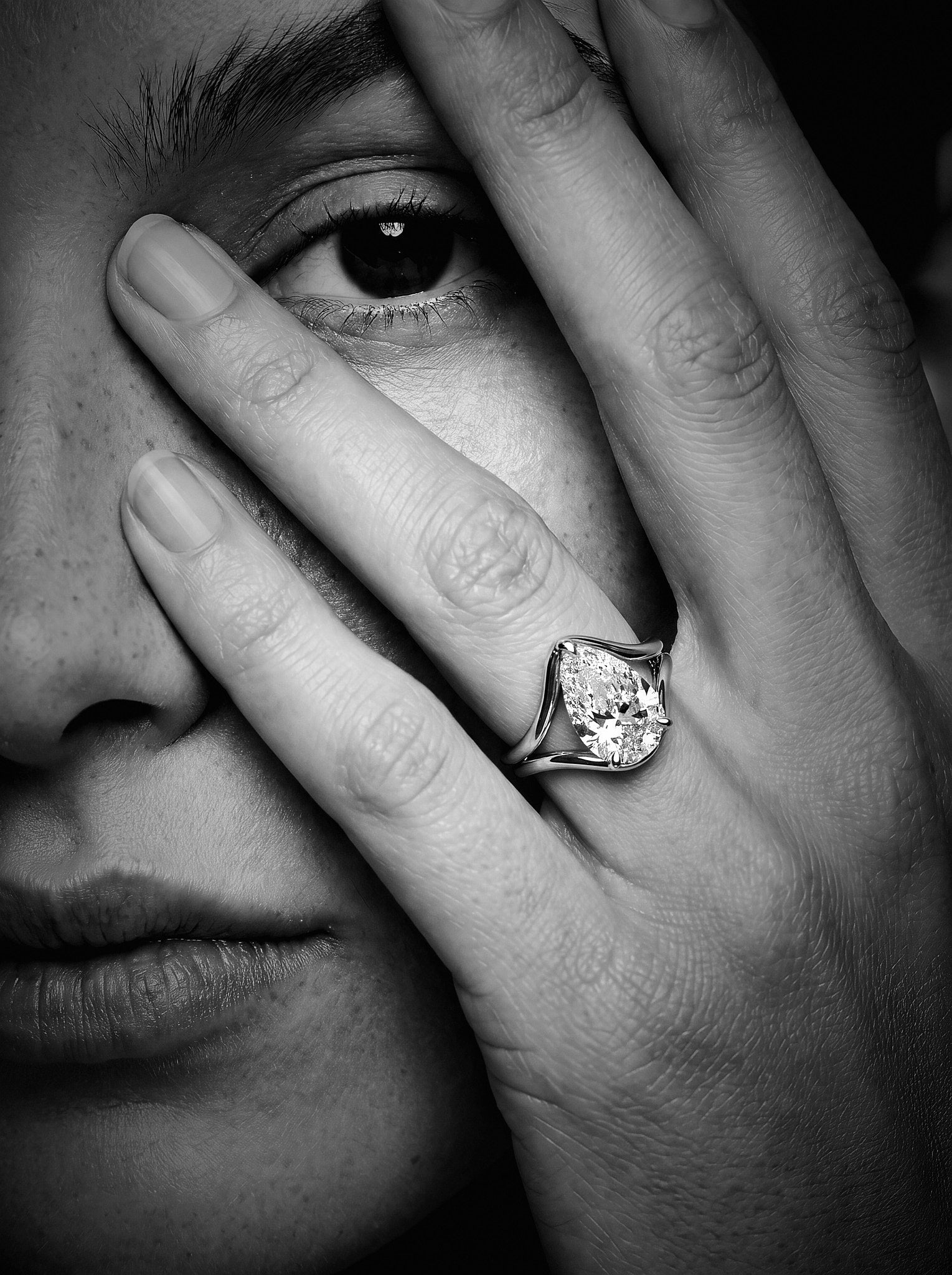 A close-up black and white photograph of a person's face and hand, with a large pear-cut diamond ring on the ring finger.