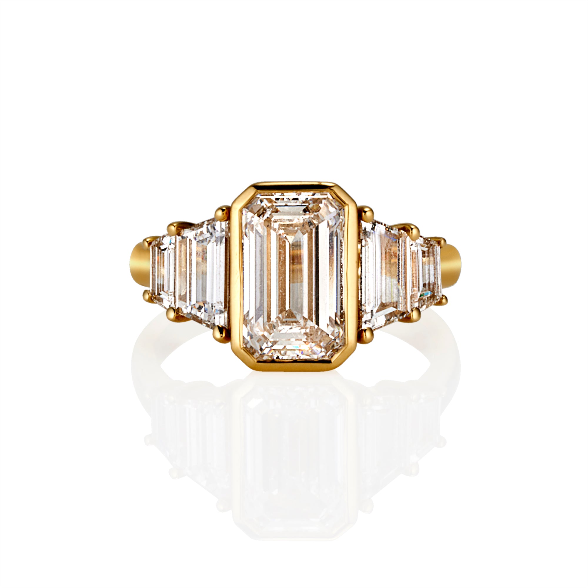 A yellow gold ring featuring a large emerald-cut clear center stone flanked by four smaller trapezoid-cut side stones, shown on a clean white background with a soft reflection below.