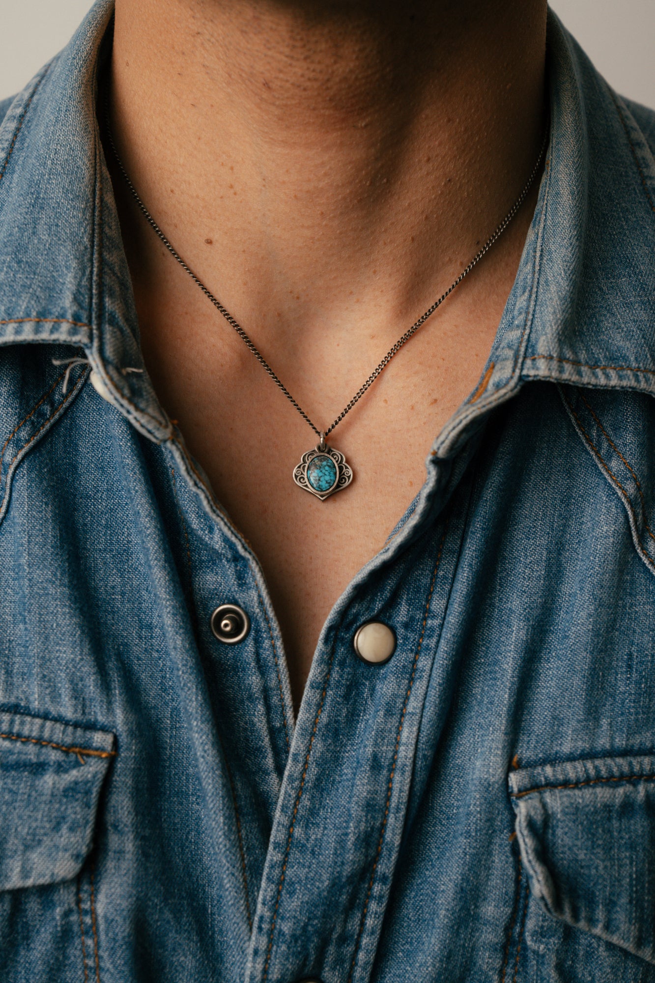 Person wearing a blue denim shirt with a necklace featuring a turquoise pendant.