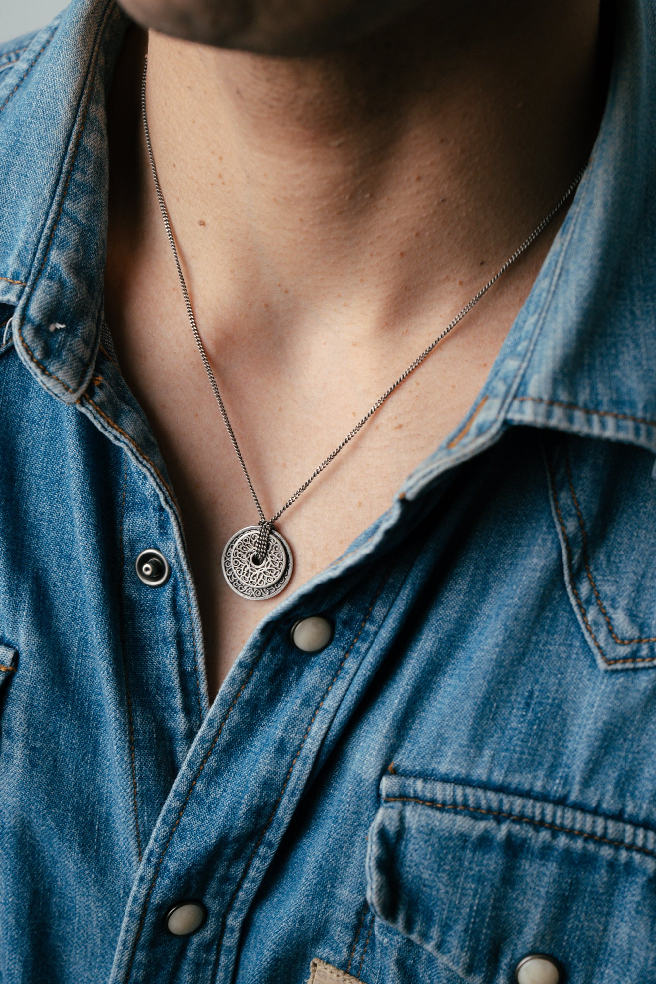 Person wearing a blue denim shirt with a necklace featuring a circular pendant.