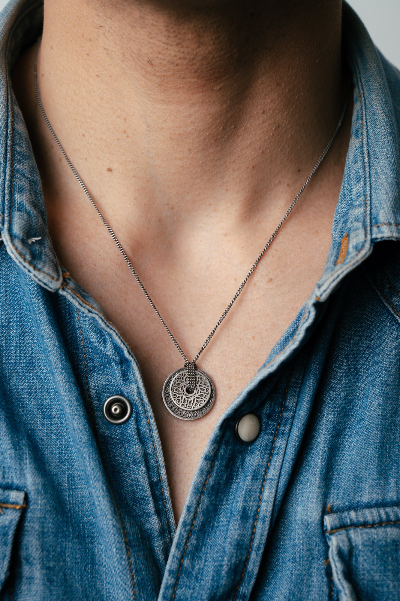 Person wearing a blue denim shirt with a necklace featuring a circular pendant.