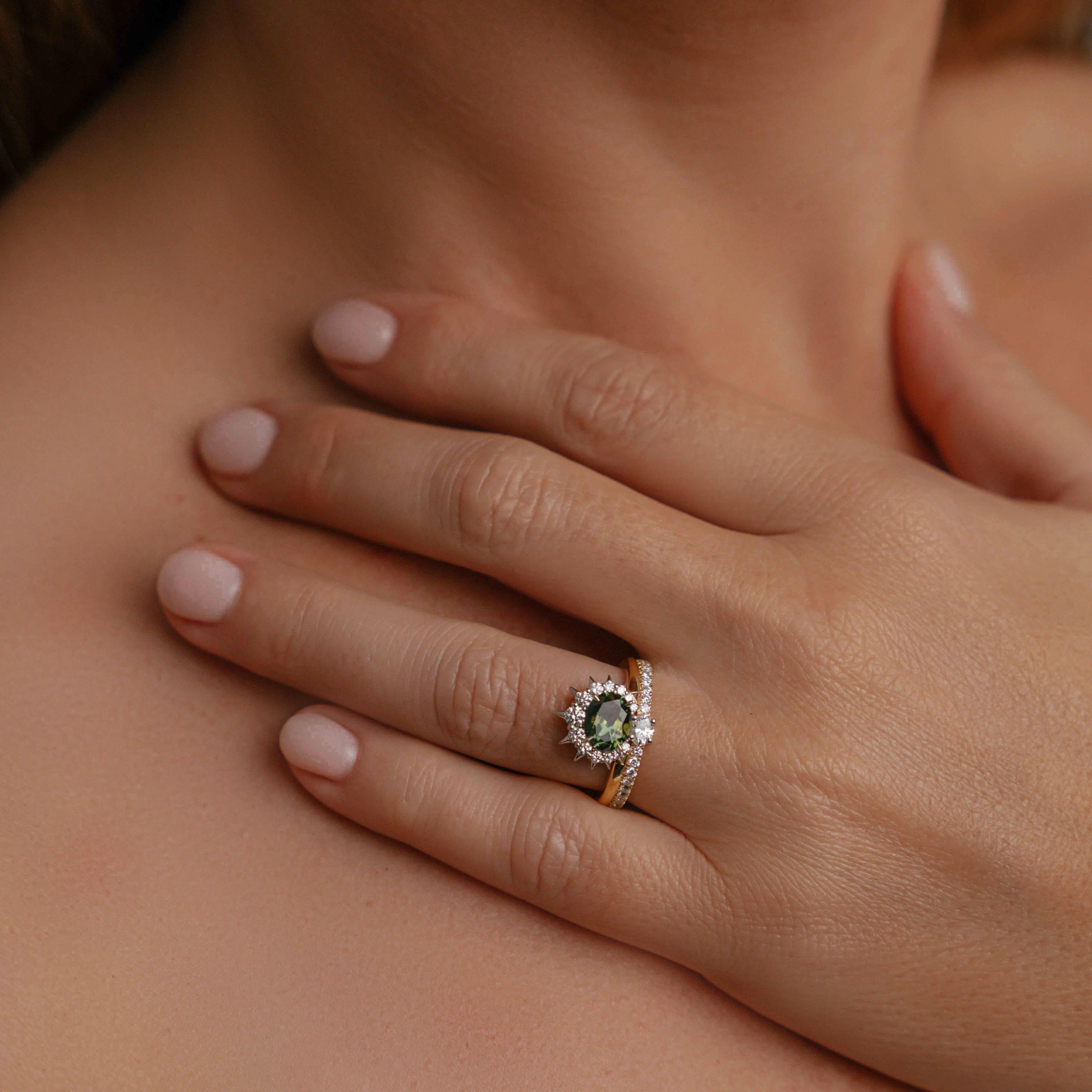 Model wearing ring on engagement ring finger, showcasing the 18ct gold & platinum engagement ring with an oval blue-green Australian sapphire, spiked diamond halo, and smaller diamonds