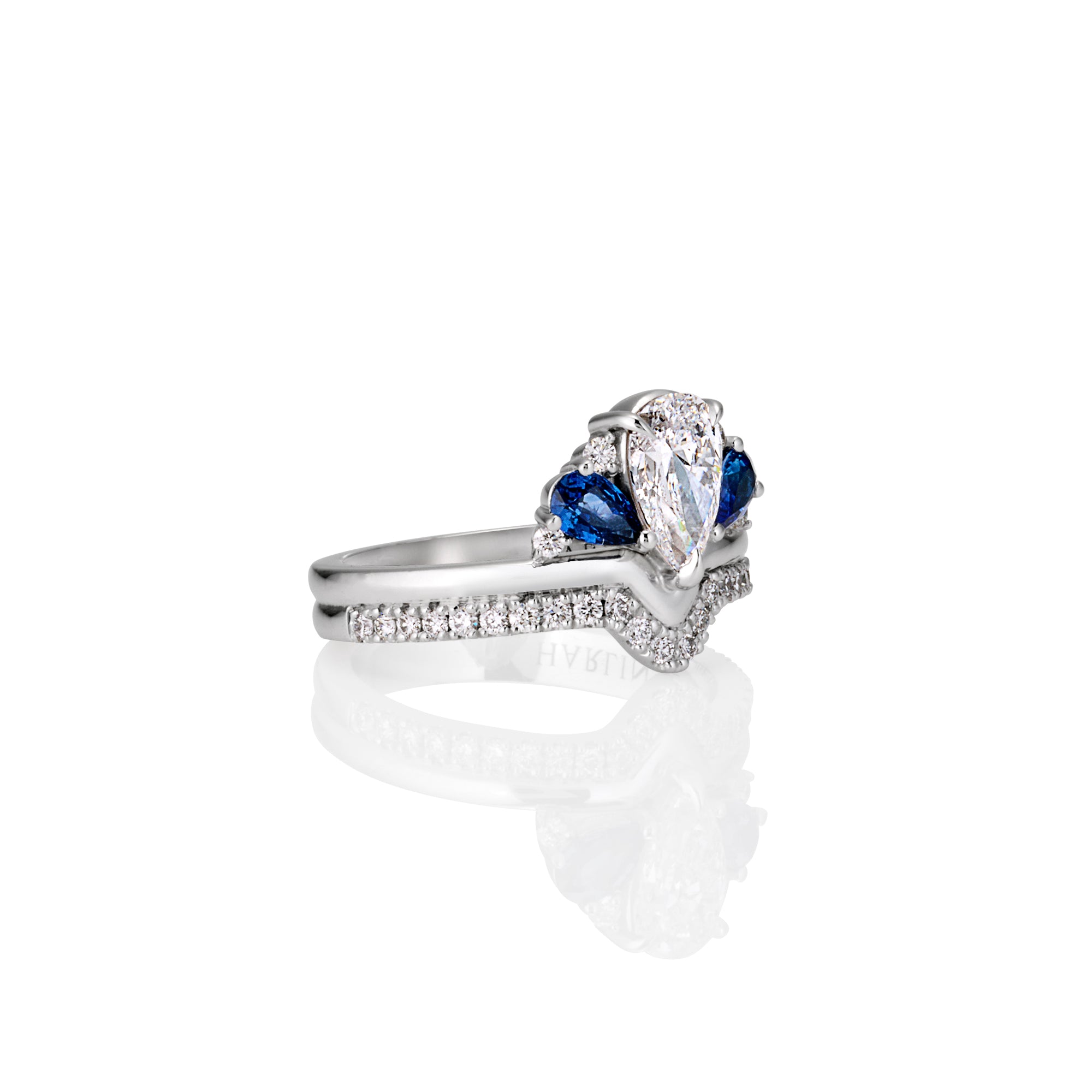 Side angle of  ring white gold with a central large pear-shaped white diamond, flanked by  blue sapphires, and a band encrusted with small sparkling diamond