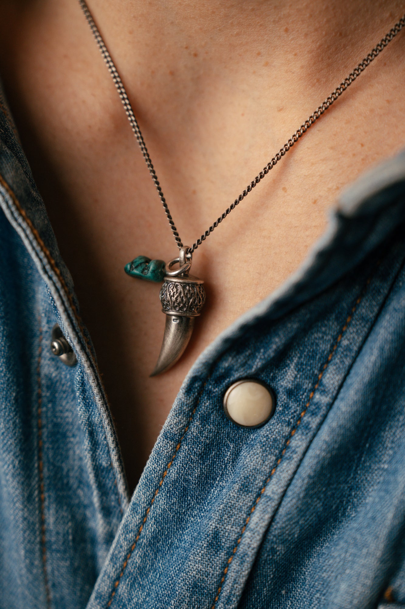 Necklace with a tusk and green stone pendant worn by a person in a denim jacket.