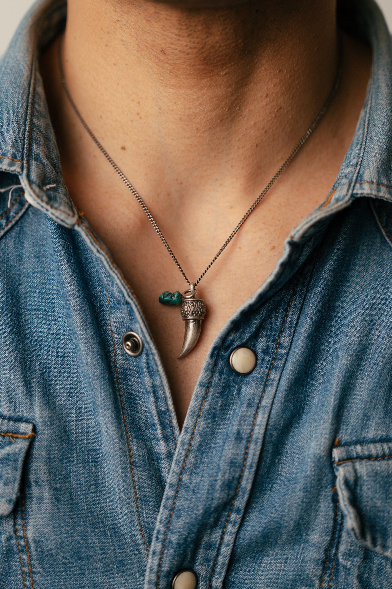 Person wearing a denim shirt with a necklace featuring a tusk pendant.