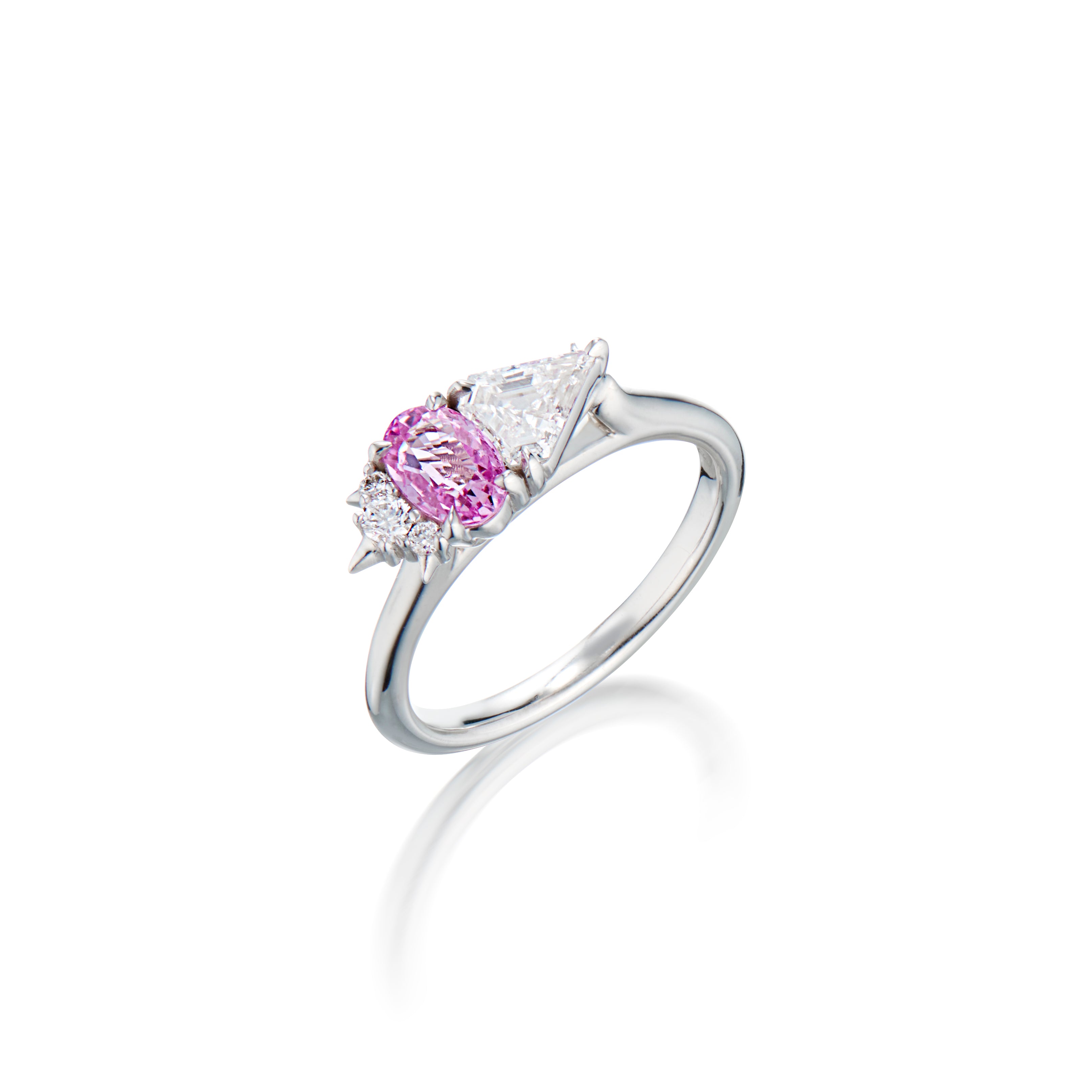 White gold ring with oval pink sapphire gemstone flanked by diamonds on each side on a reflective surface.