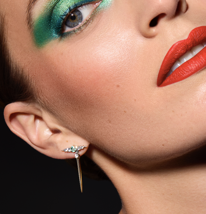 The image features a close-up of a model showcasing bold makeup, with striking green eyeshadow and vibrant red lipstick. She's wearing a pair of eye-catching earrings that feature green gemstones and diamond accents, with a distinctive spike design. The overall look combines elegance with a modern artistic flair.