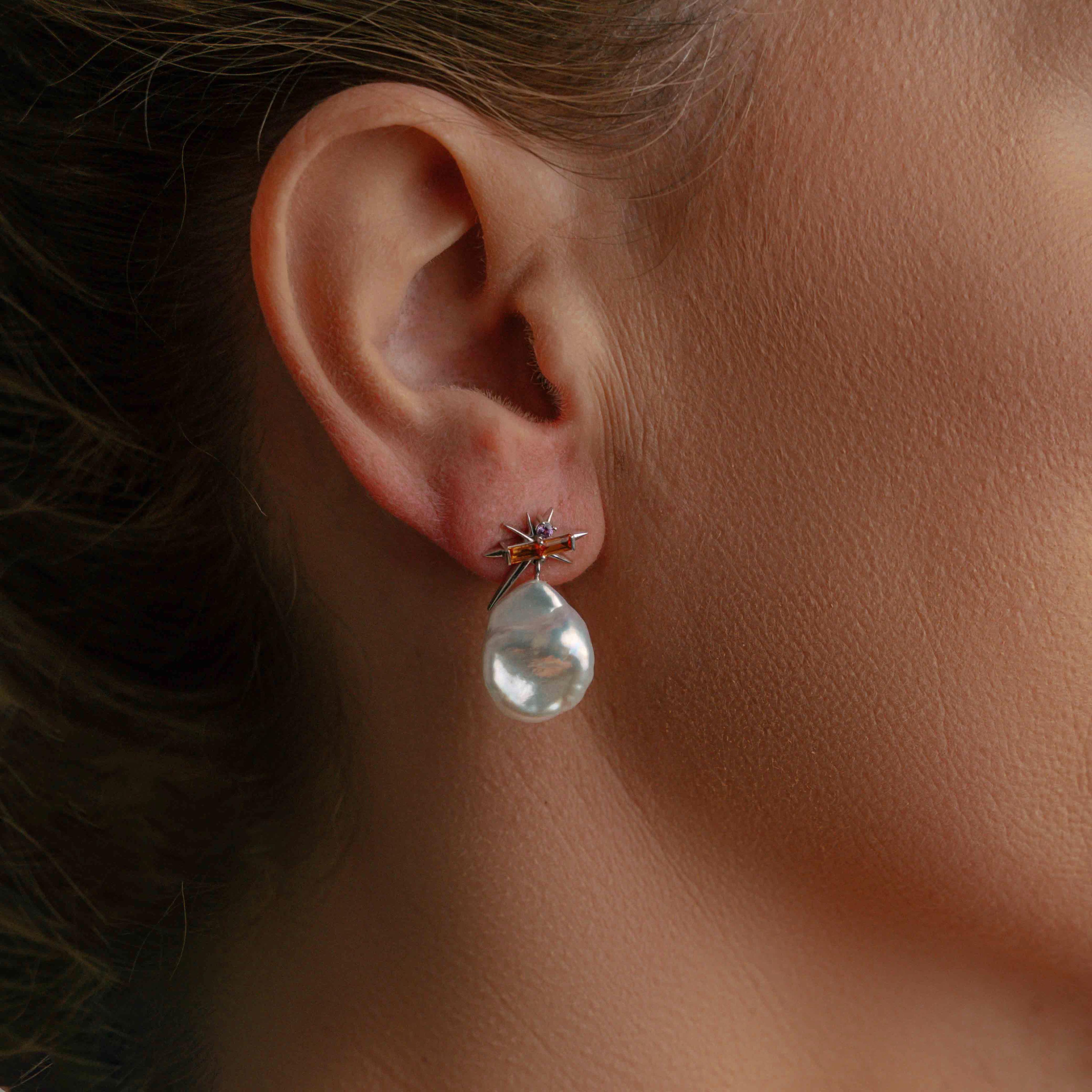 Model wearing elegant white gold earrings feature irregular white pearls, pink sapphires, and starburst accents, creating a striking and modern design.
