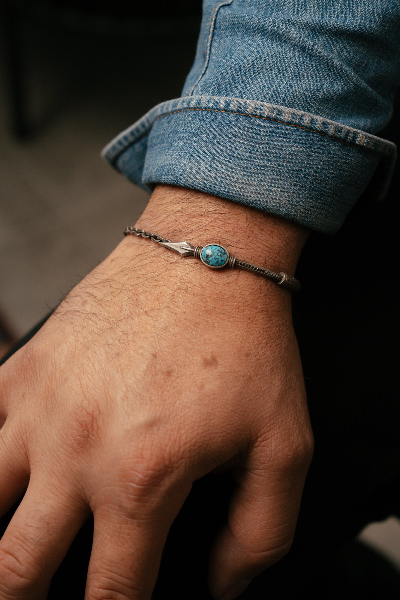 Hand wearing a bracelet with a turquoise stone, against a blurred background.