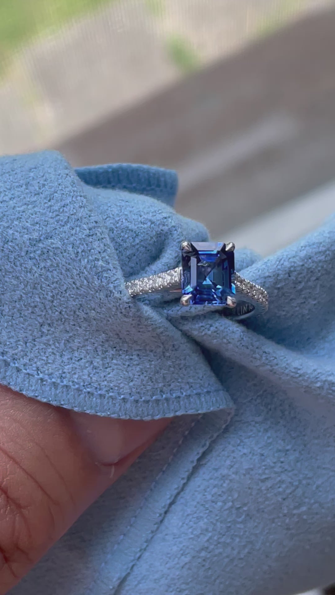 Video of white gold ring with an emerald-cut blue sapphire gemstone and diamond-encrusted band on blue cloth .