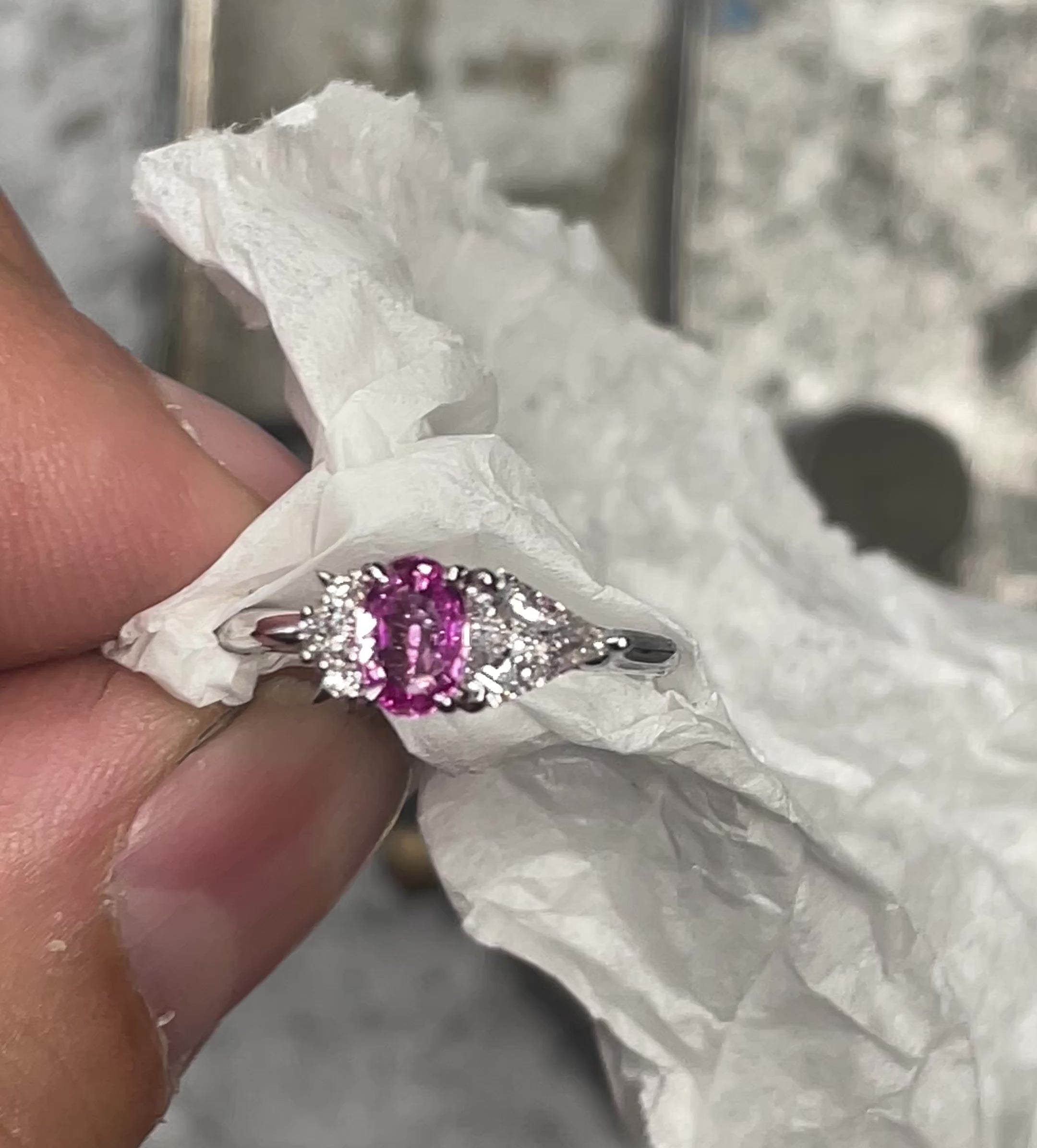 Video of White gold ring with a pink sapphire gemstone flanked by two clear diamonds, wrapped in white paper.