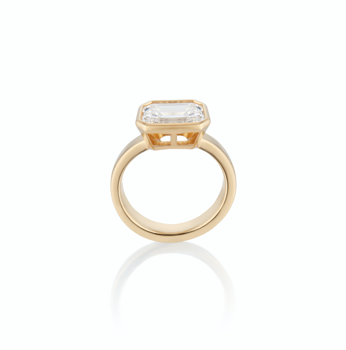 Gold ring with a single Emerald Cut diamond sitting sideways, on a white background.