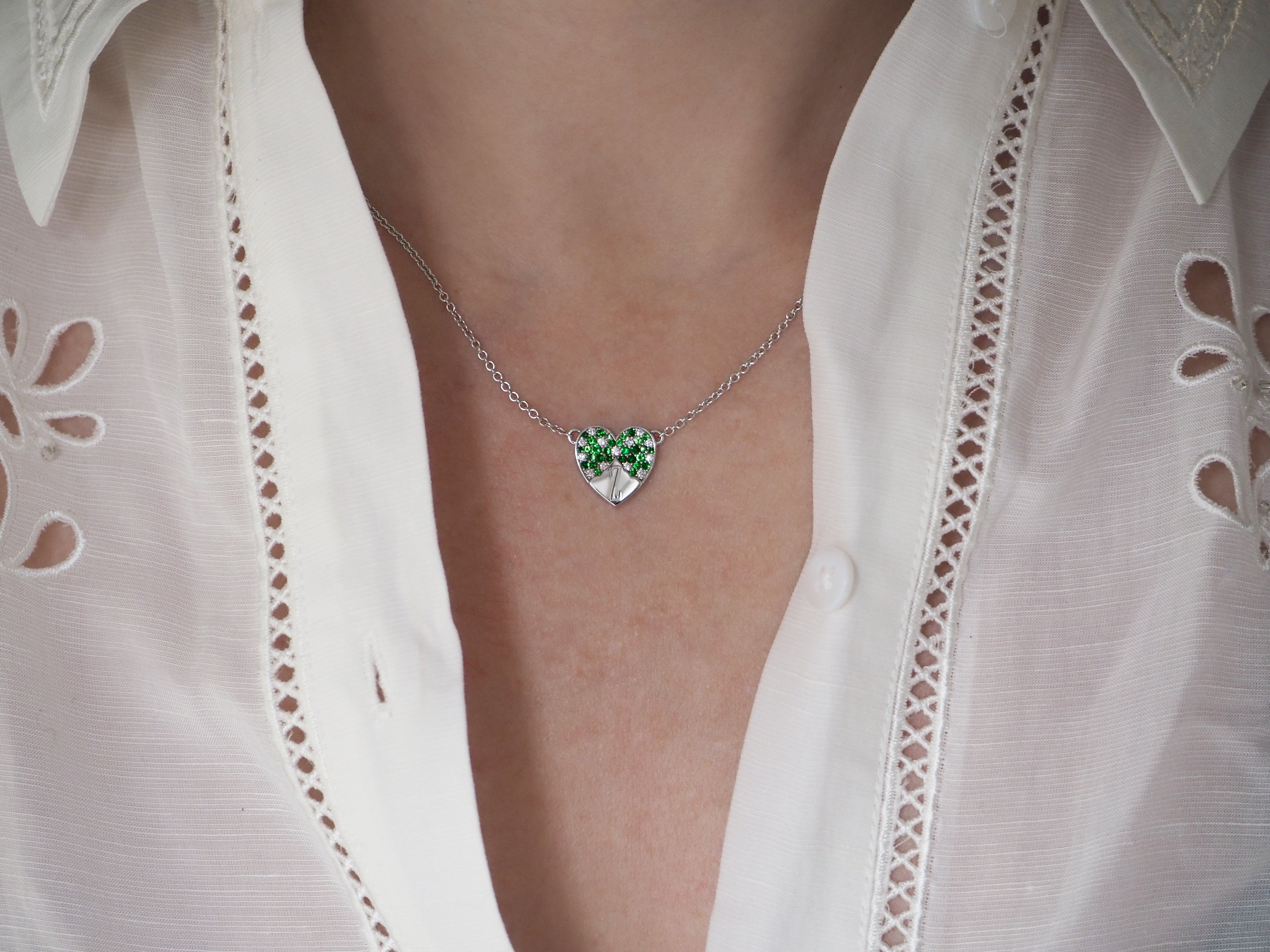 A silver necklace with a heart-shaped pendant featuring green gemstones worn with a white blouse.
