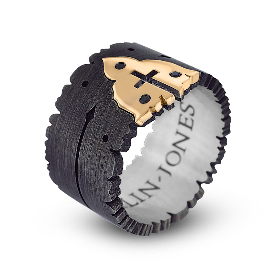 Partial side angle. Sacred Heart metal cross ring balances on base. Thick oxidized silver band with irregular jagged edges on both sides and brushed finish. The ring has a yellow gold insert in the shape of a church with 3 round black diamonds that surround a cutout shape of a cross. The interior band has an engraving that reads, “Harlin Jones”. The band’s side has an engraving of an arrow that points to the yellow gold insert.