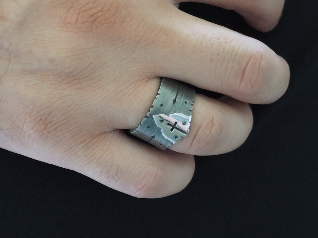 Closeup on man’s hand, which is wearing the Sacred Heart sterling silver cross ring on his ring finger. The thick band extends from the base of the knuckle to just below the first joint. The cutout cross is centered on the knuckle, with the ring’s side arrow engravings visible. The top edge of the ring comes to a point at the base of the cross.