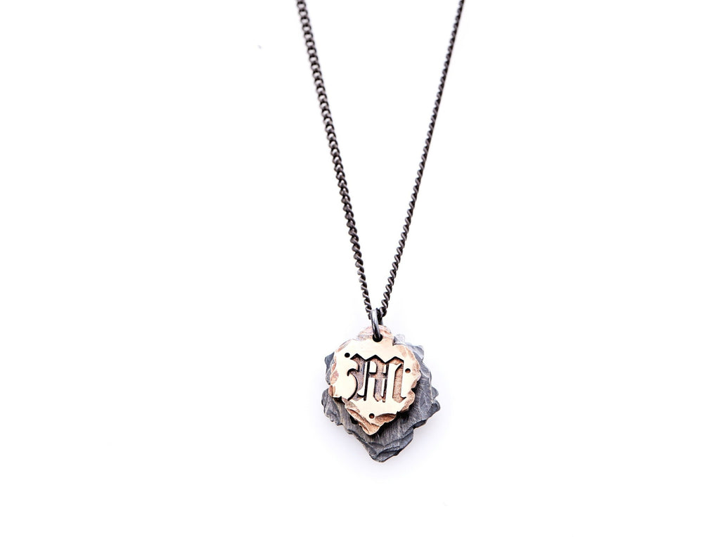 Hand crafted, customizable Initial Necklace with 2 jagged-edged plates hanging from an oxidized sterling silver curbed chain. Top plate is hand forged rose gold with a custom engraving of the letter "M" in Old English lettering with 2 hand engraved dots on either side. Bottom plate is hand forged oxidized sterling silver and slightly larger than the rose gold plate. The top plate partially covers the bottom plate, and edges of cross- bones are visible on the bottom plate.