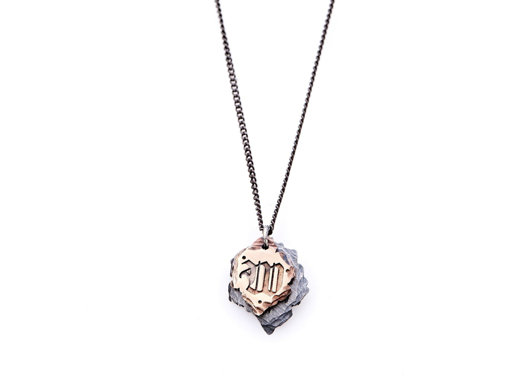 Hand crafted, customizable Initial Necklace with 2 jagged-edged plates hanging from an oxidized sterling silver curbed chain. Top plate is hand forged rose gold with a custom engraving of the letter "W" in Old English lettering with 2 hand engraved dots on either side. Bottom plate is hand forged oxidized sterling silver and slightly larger than the rose gold plate. The top plate partially covers the bottom plate, and edges of cross- bones are visible on the bottom plate.