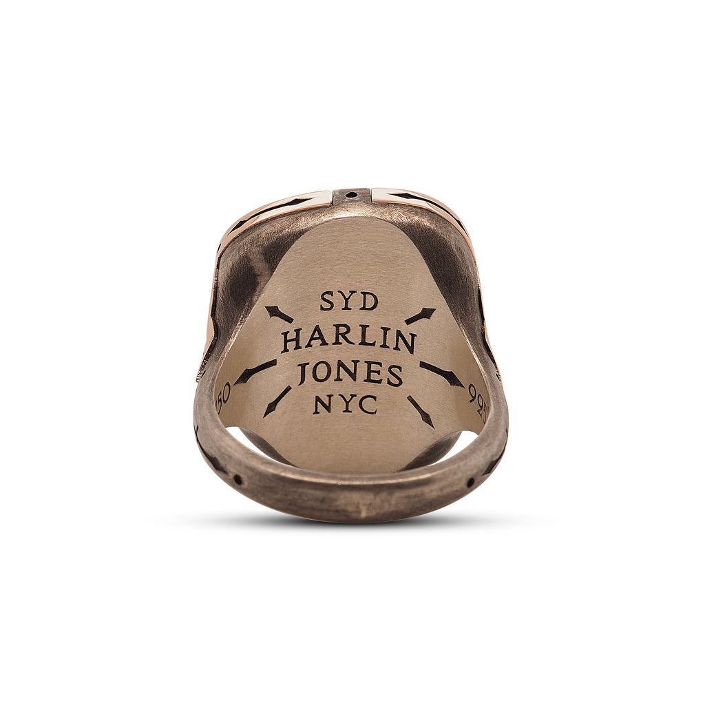 Back view of the turquoise ring, behind the sterling silver base. The interior of the ring’s head has an engraved logo that reads, “SYD Harlin Jones NYC” with six arrows pointing outwards from the logo. The left arrow points to an engraving of the number 750 and the right arrow points to an engraving of the number 925. The top of the ring is oblong, providing a unique rounded square shape.