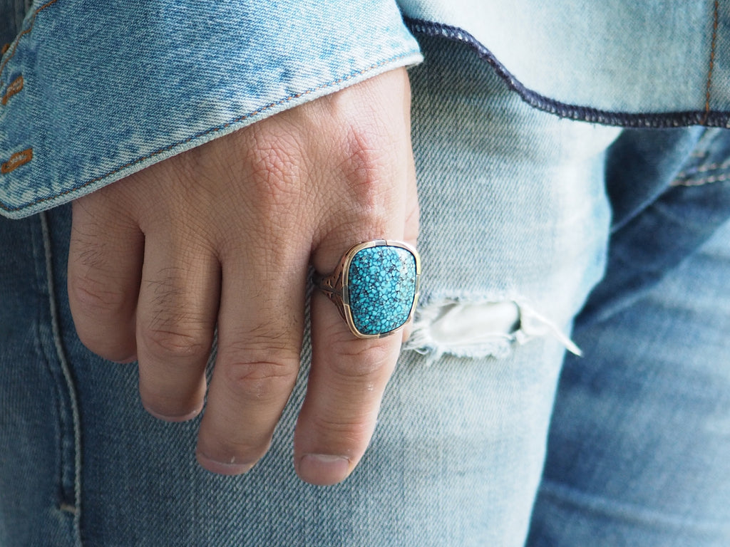 Closeup on man’s finger wearing the Kingman Turquoise Ring on his forefinger. The thin band wraps around the knuckle. The stone extends from the base of the knuckle to the first joint of the finger. Rounded square shape of the ring’s head sits flat against the finger.