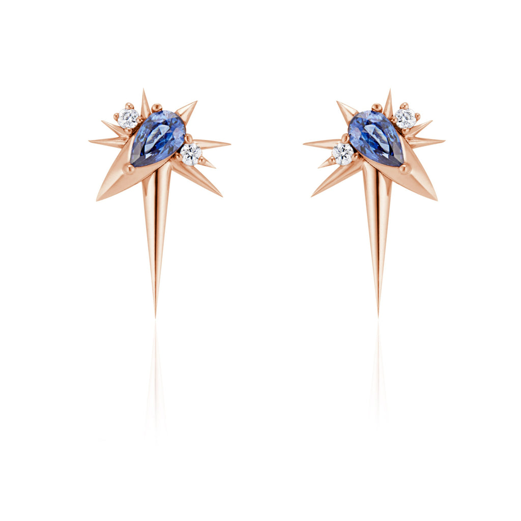 Front view of slanted drop earrings, each with 1 pear-shaped blue sapphire, set at 45-degree angle, flanked by small round diamonds. 9 rose gold spikes of varying lengths extend from the oval setting, with a larger and longer spike pointed down.
