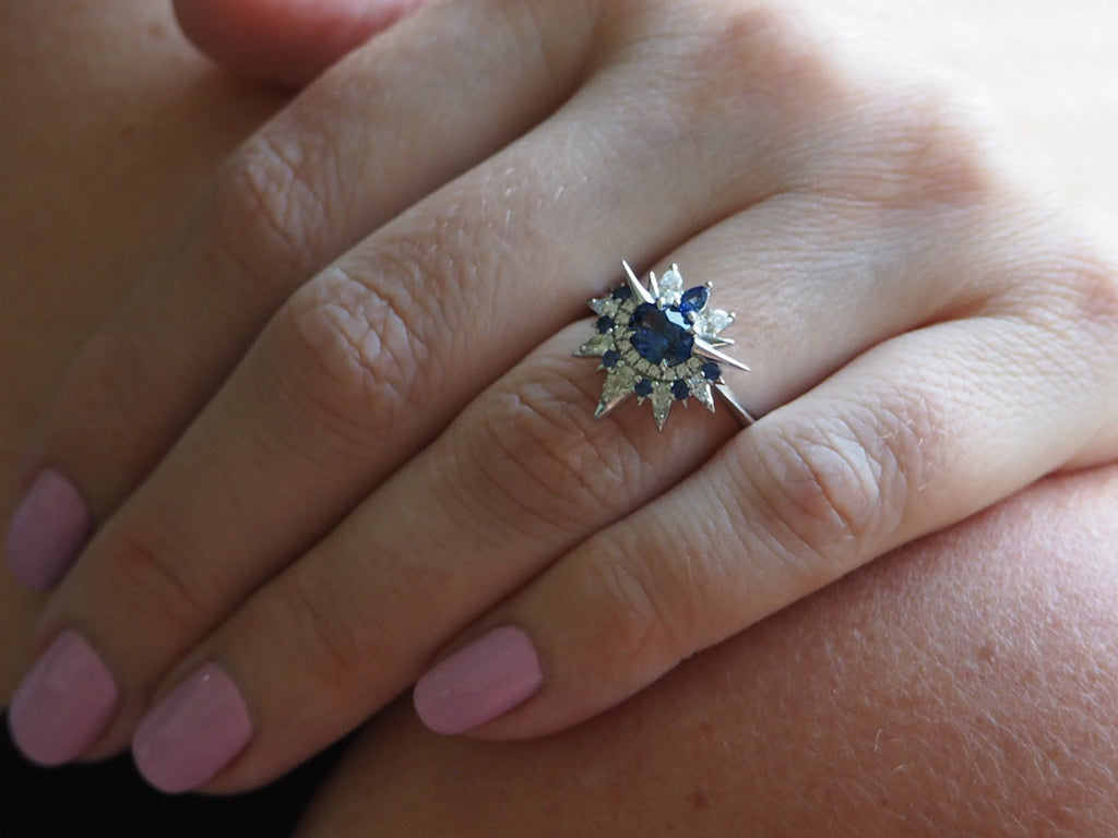 Closeup on woman’s hand wearing the blue sapphire and diamond shooting star ring on her ring finger. The thin band sits around her knuckle, just below the first joint. The ring’s head has a large round sapphire surrounded by a halo of diamonds and smaller sapphires that burst outwards in the shape of a star.