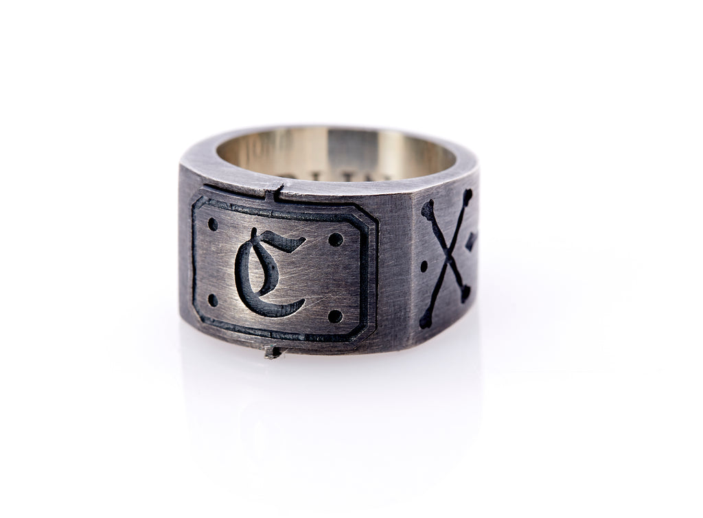 Oxidized sterling silver men’s signet ring with a thick flat band hand-crafted in New York. Semi-raised octagonal engraved top cap with a customizable insignia design of an engraved letter “C” in Old English Style lettering and 4 engraved dots at each corner. The initial on the ring is surrounded by a bold black engraved border that follows the inside edge of the rectangle. The rectangle’s top and bottom edge is accented by a small lip in the center that wraps around the corner edge of the ring.