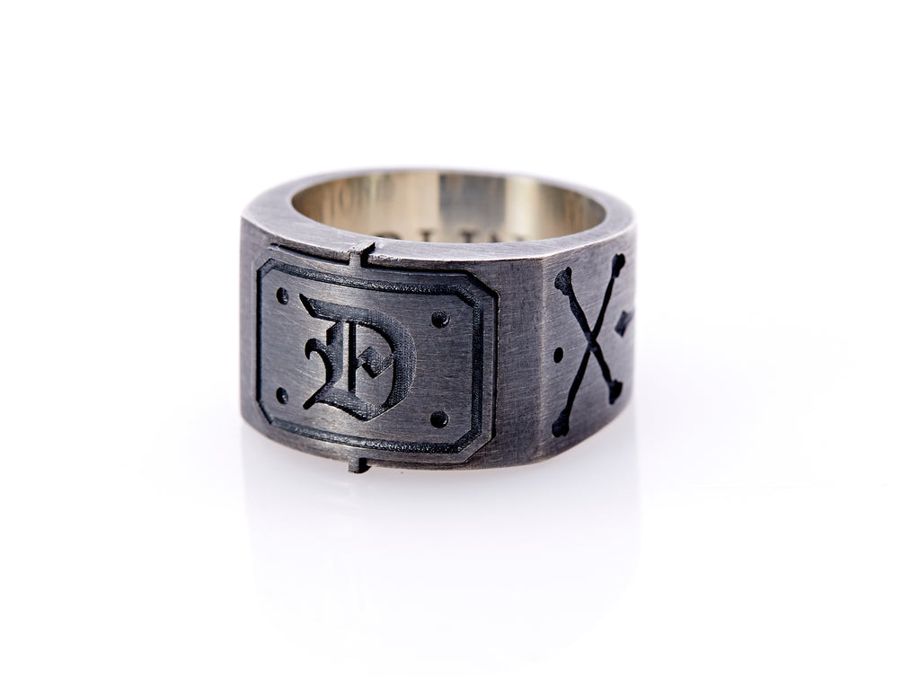 Oxidized sterling silver men’s signet ring with a thick flat band hand-crafted in New York. Semi-raised octagonal engraved top cap with a customizable insignia design of an engraved letter “D” in Old English Style lettering and 4 engraved dots at each corner. The initial on the ring is surrounded by a bold black engraved border that follows the inside edge of the rectangle. The rectangle’s top and bottom edge is accented by a small lip in the center that wraps around the corner edge of the ring.